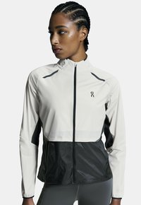 Chaqueta ligera con una sección superior color crema y la mitad inferior negra, con cierre de cremallera, cuello alto y tejido transpirable.