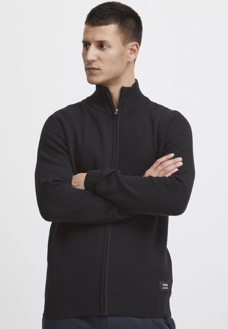 Svart zip-up tröja med hög krage, stickad textur, ribbade muddar och kant. Har en liten varumärkesetikett på nedre vänstra sidan.