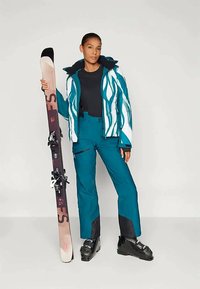 Síelő öltözet, amely egy türkiszkék dzsekit tartalmaz fehér hullámos mintákkal, fekete thermál pólót, cipzáras türkiszkék nadrágot és fekete síbakancsot, miközben rózsaszín síléceket tart.