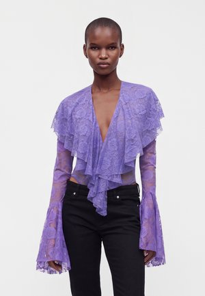 Personne portant un chemisier en dentelle violette transparente avec des volants superposés et des manches évasées, associé à un pantalon noir sur un fond uni.