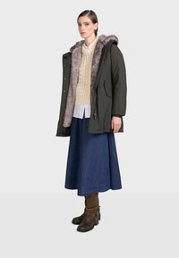 Parka w ciemnozielonym kolorze z futrzanym wykończeniem, zestawiony z kremowym swetrem w warkocze, dżinsową spódnicą i brązowymi botkami na obcasie z paskami.