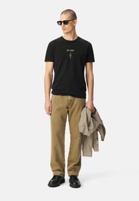 Camiseta negra de algodón con impresión "EN ESCENA", pantalones vaqueros de pana color beige, zapatos negros gruesos y una chaqueta de cuadros beige colgada del brazo.