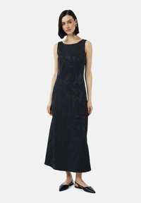 Mouwloze jurk van midi-lengte in donker marineblauwe stof met een subtiel patroon, met een getailleerde bovenkant en een wijd uitlopende onderkant. Gemaakt met zwarte slingback-schoenen.