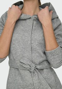 Manteau gris à capuche en tissu texturé, avec une taille ceinturée et un col à rabat. Manches longues avec des poignets retroussés.