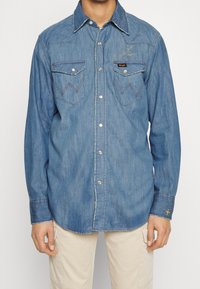 Uomo che indossa una camicia in denim blu a maniche lunghe con bottoni a pressione e due tasche a soffietto frontali, abbinata a pantaloni beige chiaro.