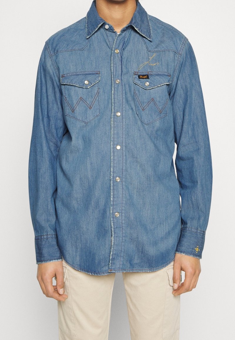 Uomo che indossa una camicia in denim blu a maniche lunghe con bottoni a pressione e due tasche a soffietto frontali, abbinata a pantaloni beige chiaro.