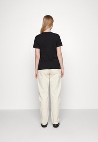 T-shirt preto de manga curta com decote redondo, usado com calças largas de cor creme e sapatos pretos, visto de costas.