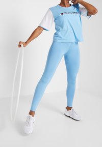 Světle modrá košile s krátkým rukávem a bílými akcenty a logem, spárovaná s odpovídajícími těsnými světle modrými legínami a bílými sportovními botami.