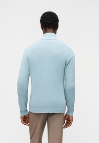 Άνδρας που φοράει ανοιχτό γαλάζιο πλεκτό πουλόβερ και καφέ παντελόνι, γυρισμένος προς τα πίσω με φόντο έναν απλό λευκό τοίχο.