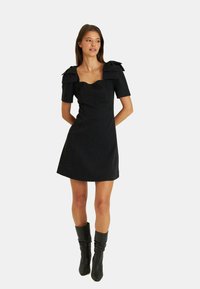 Schwarzes Kleid mit kurzen Ärmeln und Schleifenakzenten, taillierte Silhouette, strukturierter Stoff, knielanger Saum, kombiniert mit kniehohen schwarzen Stiefeln.