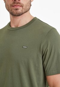 Olive green cotton T-shirt with a round neckline and short sleeves. Features a small black fabric label with "TOG24" on the chest.