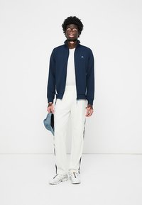 Mornarska zip-up jakna, bela majica, bele hlače z modrimi stranskimi črtami in bele superge. Model drži modro denim kapo.