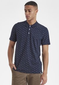 Polo de color navy con un pequeño patrón geométrico blanco, tapeta con tres botones y cuello clásico. Textura suave, mangas cortas y corte casual.