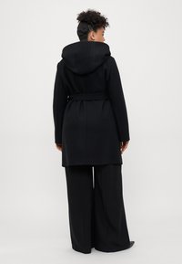 Donna con capelli ricci, che indossa un cappotto nero con cappuccio e cintura e pantaloni neri a gamba larga, rivolta verso l'esterno contro uno sfondo chiaro e semplice.