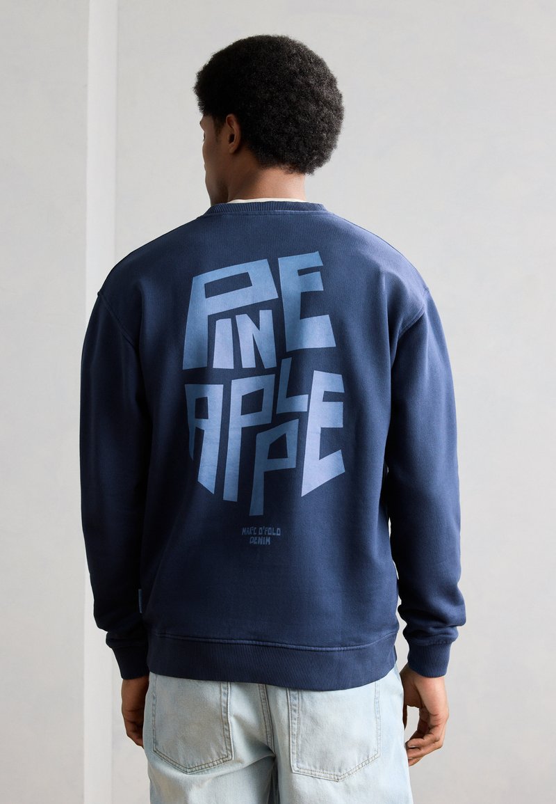 Άνδρας στραμμένος με την πλάτη, φοράει μπλε σκούρο φούτερ με μεγάλο μπλε γεωμετρικό κείμενο που γράφει «Pine in a Plape» και ανοιχτόχρωμο τζιν.