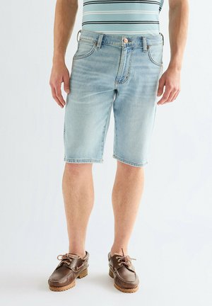 Mann, der hellblaue Jeansshorts, ein gestreiftes Hemd und braune Leder-Schnürschuhe trägt und vor weißem Hintergrund steht.