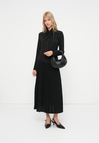 Blonda sieviete, kas valkā melnu garu piedurkņu topu un krokotu midi svārku, tur melnu rokassomu un valkā melnas atvērtās papēžsandaļas.