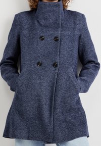 Marineblaue Jacke mit hohem Kragen, doppelreihiger Knopfleiste, zwei Fronttaschen und strukturiertem Stoff mit einem subtilen Muster.