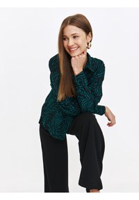 Koszula z długim rękawem zapinana na guziki w ciemnozielonym kolorze z czarnym wzorem liści. Połączona z czarnymi, luźnymi spodniami. Gładka tekstura materiału.