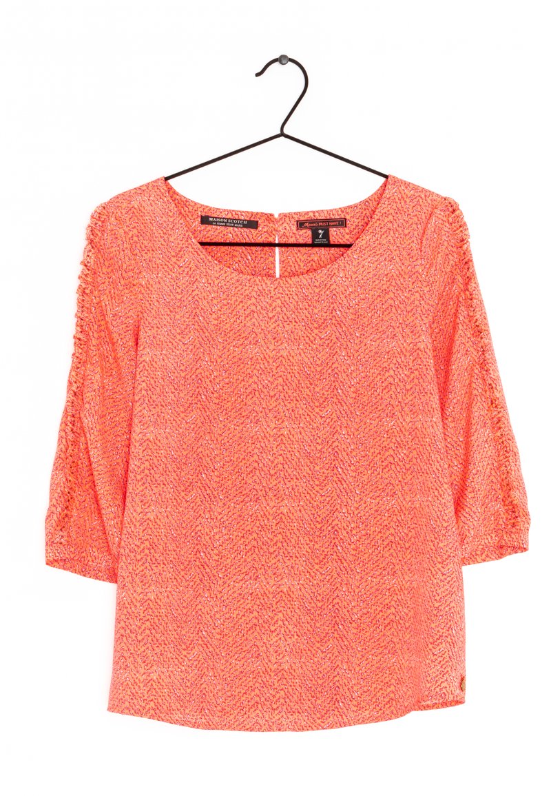 Blouse en corail texturé avec des manches bouffantes 3/4, un col rond et un subtil motif en chevrons. Suspendue à un crochet noir.
