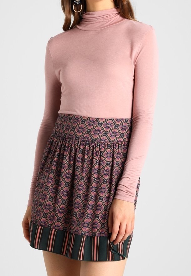 Rosa Langarm-Mockturtleneck-Oberteil, kombiniert mit einem gemusterten, gerafften Rock, der geometrische Designs in Lila und Grün mit gestreiften Akzenten aufweist.