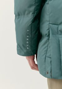 Zelená bunda s kapucňou má hladký matný povrch, postranné vrecká a text "HYDRE-T" vytlačený vertikálne na rukáve. Viditeľné textúrované švy.