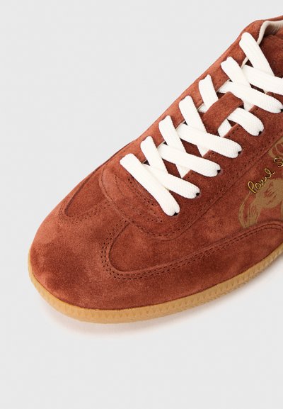 Pruun nahkahiivatud sneaker valgete paelte ja kummist tallaga, millel on kuldne kiri ja diskreetne graafika küljel.