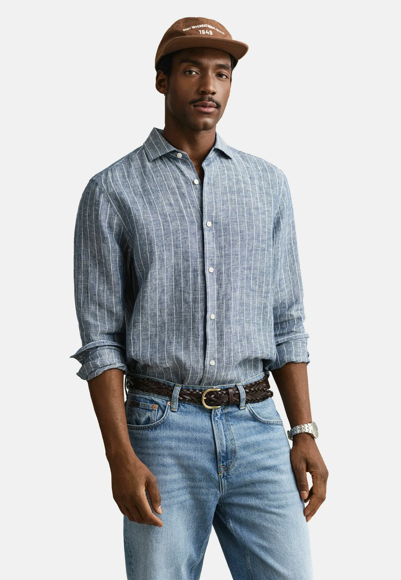 Homme portant une casquette marron, une chemise à rayures bleues et blanches à boutons avec manches retroussées, une ceinture tressée marron, un jean bleu clair et une montre-bracelet argentée.