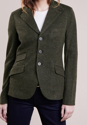 Kobieta ubrana w dopasowaną, ciemnozieloną, wełnianą marynarkę w jodełkę z trzema guzikami, klapowanymi kieszeniami i białą koszulą pod spodem.