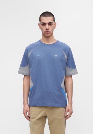Giovane uomo con capelli corti che indossa una maglietta sportiva a maniche corte blu e grigia e pantaloni beige, in piedi davanti a uno sfondo bianco uniforme.
