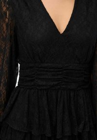 Robe en dentelle noire avec manches longues transparentes, décolleté plongeant en V, taille froncée et jupe à volants étagée.