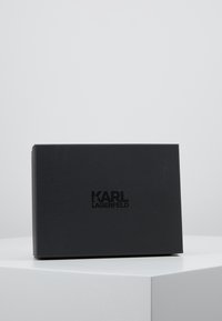 Czarny pojemnik o gładkim, matowym wykończeniu, z wytłoczonym napisem "KARL LAGERFELD" z przodu. Prosty prostokątny kształt z czystymi krawędziami.