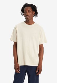 T-shirt en coton beige clair avec un col ras du cou et des manches courtes. Présente un détail de logo subtil sur la poitrine ; coupe décontractée et texture douce.