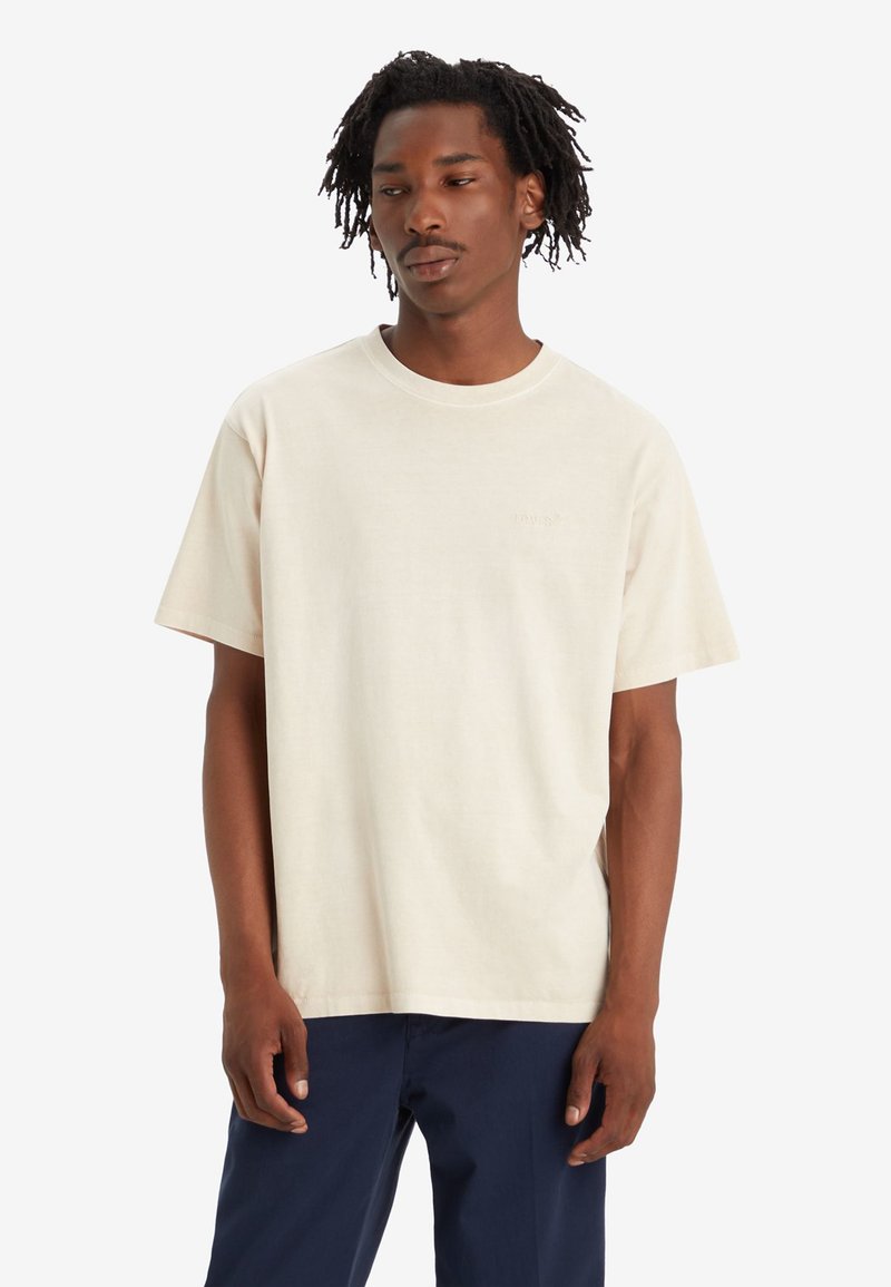 T-shirt en coton beige clair avec un col ras du cou et des manches courtes. Présente un détail de logo subtil sur la poitrine ; coupe décontractée et texture douce.