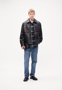 Black leather jacket with two chest pockets over a blue plaid shirt, paired with light blue jeans and black shoes. Model standing against a white background.