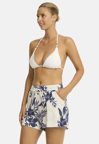 Een glimlachende vrouw poseert in een witte bikini en bloemen short, en toont een ontspannen zomerstijl tegen een effen achtergrond.