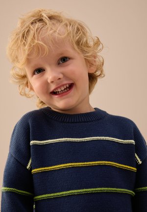 Smaidošs jauns bērns ar cirtainiem blondiem matiem, valkā tumši zilu džemperi ar dzeltenām, zaļām un baltām horizontālām svītrām.