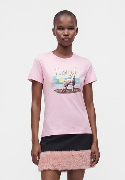 Γυναίκα που φοράει ανοιχτό ροζ T-shirt με ένα αρνάκι και τη λέξη "Rebel,", σε συνδυασμό με μια μαύρη φούστα με ροζ τεχνητή γούνα στο τελείωμα.