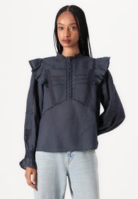 Dark navy blouse with ruffled shoulders, high neck, buttoned front, and embroidered detailing. Long sleeves with elastic cuffs and textured fabric.