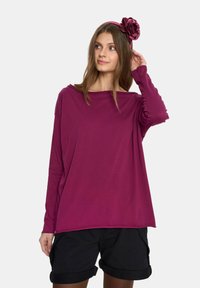 Różowa bluzka z długim rękawem uszyta z miękkiego materiału, z luźnym krojem i szerokim dekoltem, połączona z czarnymi szortami z podwiniętymi nogawkami oraz dopasowaną opaską na głowę.