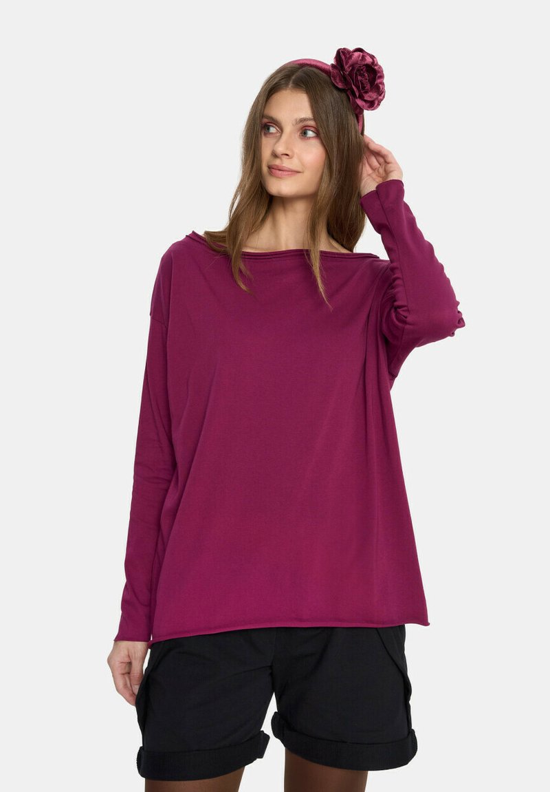 Różowa bluzka z długim rękawem uszyta z miękkiego materiału, z luźnym krojem i szerokim dekoltem, połączona z czarnymi szortami z podwiniętymi nogawkami oraz dopasowaną opaską na głowę.