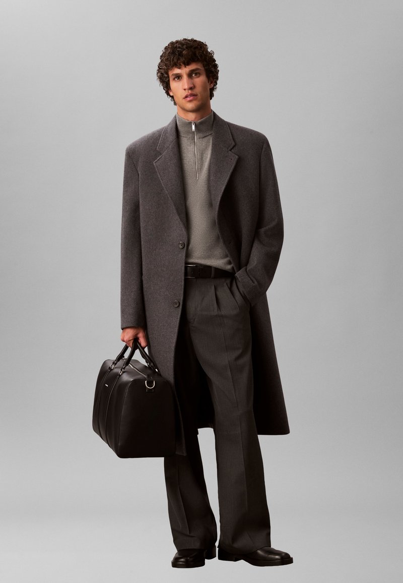 Man wearing a long gray overcoat, gray zip-up sweater, dark trousers, black shoes, holding a black leather briefcase against a plain background.