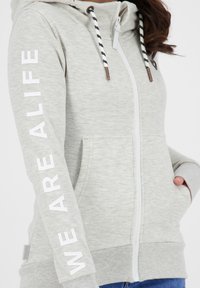 Jasnoszara bluza z kapturem na zamek, wykonana z miękkiej tkaniny, z kieszenią typu kangurek, ozdobiona nadrukiem "WE ARE ALIFE" na rękawie oraz paskowanymi sznurkami.