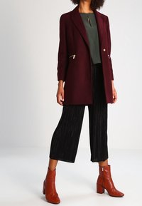 Kvinna som bär en vinröd kappa över en grön topp, svarta plisserade vida byxor och bruna ankelnaglar med blockklackar.