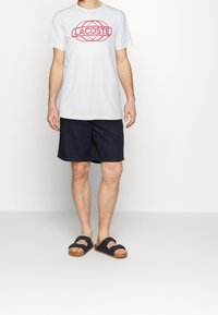 Bílý bavlněný tričko s červeným logem Lacoste, doplněné modrými kraťasy a černými sandály s korkovou podrážkou. Jednoduchý design, volný střih.