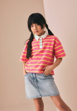 Ung pige med langt mørkt hår i to fletninger iført en polo med lyserøde og gule striber og en lyseblå denim nederdel, hænderne i lommerne.