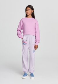 Felpa cropped rosa chiaro con maniche lunghe abbinata a pantaloni tuta lavanda con grafiche e motivi colorati. Sneakers blu completano il look.