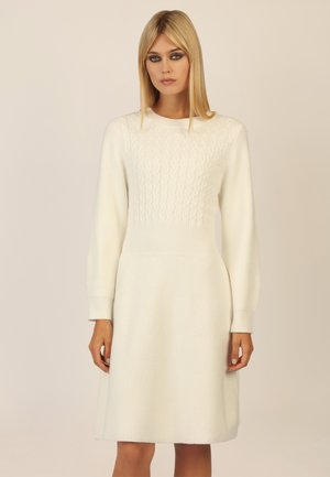 Weißes gestricktes Kleid mit einem strukturierten Zopfdesign im Oberteil und gerippten langen Ärmeln, die zu einem ausgestellten Rock verjüngen. Weiche, warme Textur.