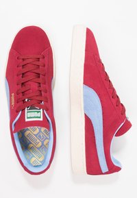 Röda mockaskor med blå accenter, med en strukturerad yta, platta snören och en krämfärgad gummisula. PUMA-logotyp synlig på tungan.