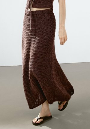 Femme marchant dans une jupe maxi marron en crochet et un haut assorti, portant des sandales tongs noires sur un sol uni.